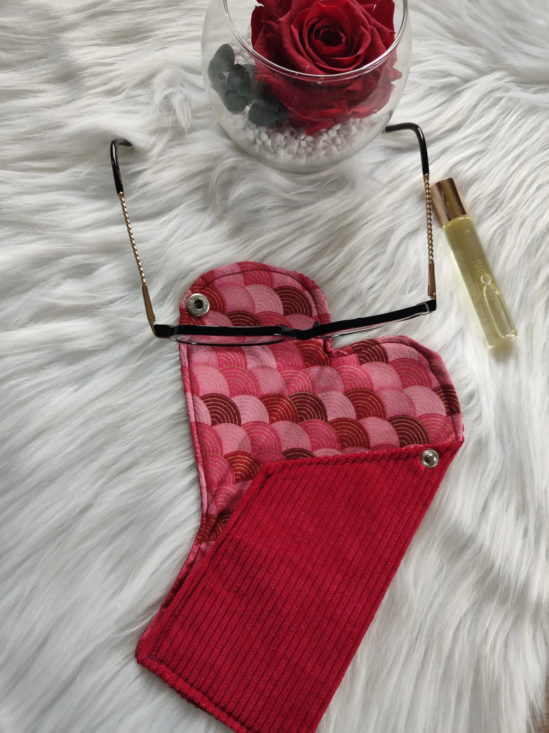 Etui à lunette confectionner avec un tissu côtelé rouge pour l'extérieur et un tissu avec des forme géométrique de couleur rouge, rose et doré. La photo est composé également d'une paire de lunettes, d'un petit tube de parfum et d'un pot en verre avec une rose rouge séché à l'intérieur.