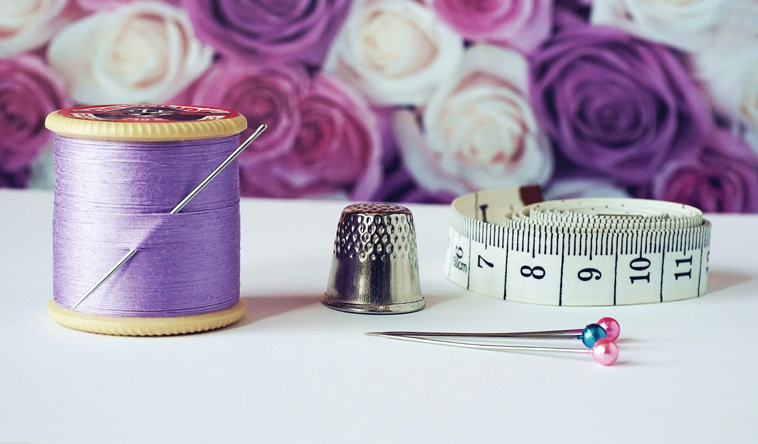fil violet ensemble de couture représentant une bobine de fil de couleur violette avec une aiguille planter dedans, un dé de couture, des épingles avec des tête de couleur, rose et bleu et un mètre de couture, le tous avec un fond de fleur de couleur violette et blanche