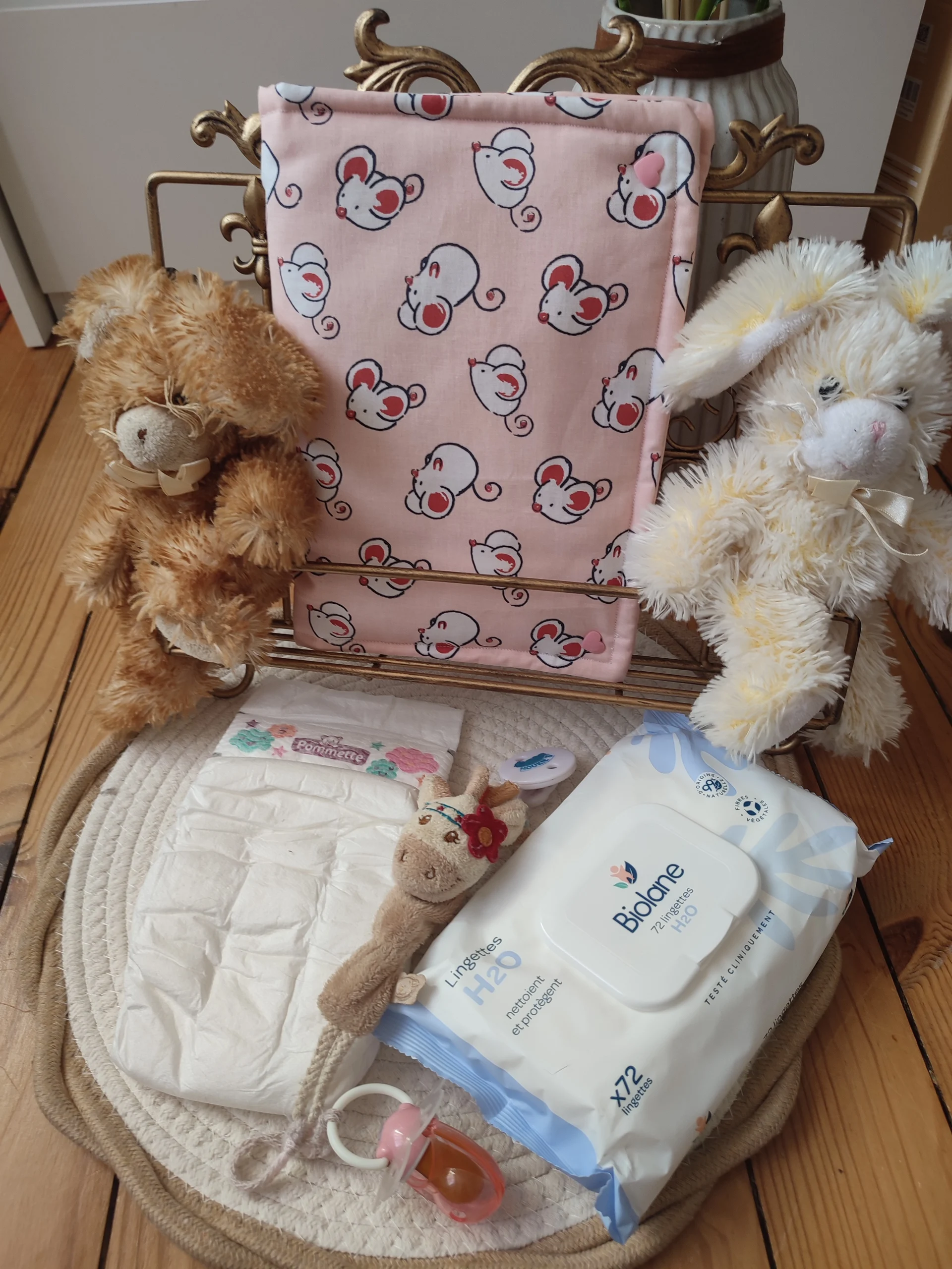 pochette à lange nomade fermé de couleur rose et avec des motifs souris sur présentoir accompagné de deux peluches lapin de couleur crème et marron, une couche, une attache tétine en forme de vache avec une tétine rose et un paquet de lingette blanche et bleu, le tous sur un tapis rond couleur beige et crème