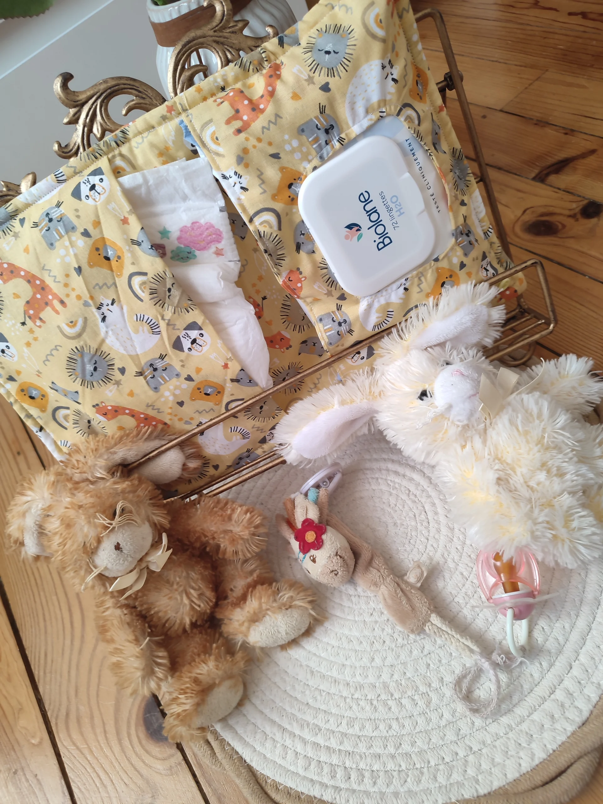 pochette à lange nomade ouverte de couleur jaune avec des motifs d'animaux sur présentoir avec à l'intérieur une couche et une paquet de lingette de couleur blanc et bleu accompagné de deux peluches lapin de couleur crème et marron, une attache tétine en forme de vache avec une tétine rose , le tous sur un tapis rond couleur beige et crème